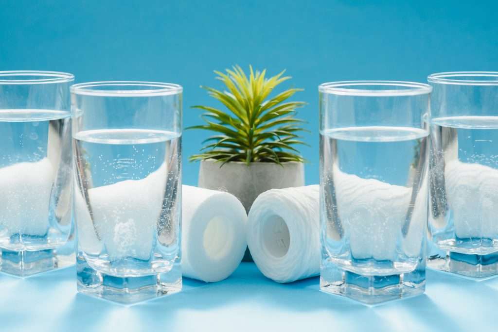Glasses of dirty and clear water and filter cartridges to domestic water treatment systems on a bright blue background.